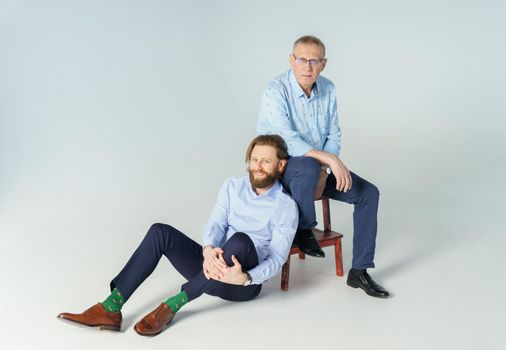 Father sits on the chair and son sits on white background, both men look into the camera, an elderly man in glasses with diopters, a stylish young man with a beard and long hair. High quality photo