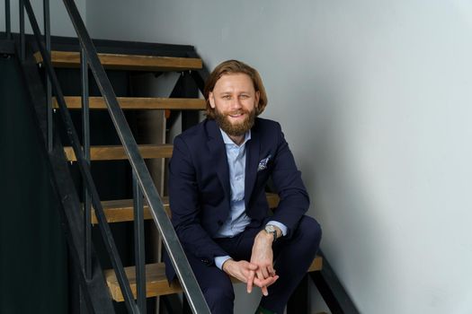 a handsome brutal bearded and long-haired man sits on the steps of the stairs, a stylish businessman. High quality photo