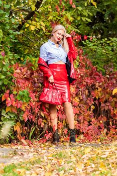 A fashionable woman wears a trendy white and blue shirt and red skirt and leather jacket on a city street. Stylish young blonde lady with bright makeup.