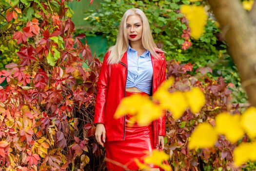 A fashionable woman wears a trendy white and blue shirt and red skirt and leather jacket on a city street. Stylish young blonde lady with bright makeup.