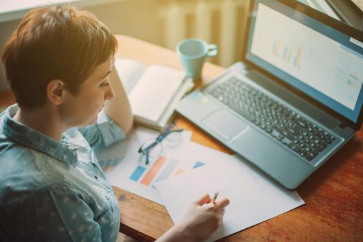 Working at home. Top view of a woman accountant analyst doing a graph analysis in business, taking notes while working from home, sitting at a table with a laptop