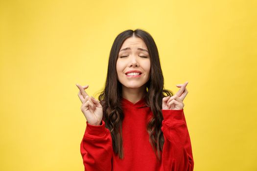 Intrigued woman in praying with crossed fingers and looking away over yellow background.