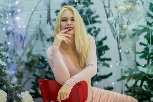 Charming blonde woman in beige sexy dress sitting on red chair, over background with Christmas tree. Christmas and New Year photo.