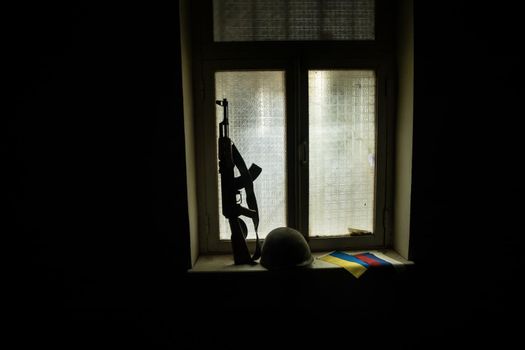 Conceptual photo of war between Russia and Ukraine. Ukrainian flag, helmet and gun on windowsill at night. Old creepy room with window. Explosion outside.