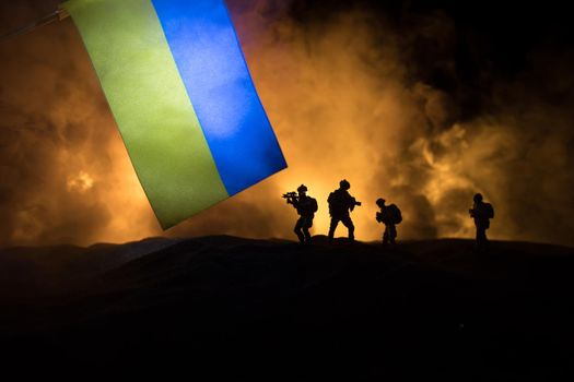 Russian war in Ukraine concept. Silhouette of armed soldiers against Ukrainian flag and burned out city. Creative artwork decoration. Night fighting scene. Selective focus