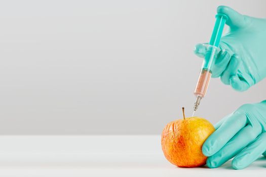 A hand in a medical glove inserts a syringe into apple. Harmful food additives. GMOs Concept. Injecting on apple at laboratory. Fruit genetic modification concept.