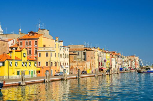 Narrow water canal with promenade embankment and colorful buildings in Chioggia town historical centre, blue sky background in summer day, Veneto Region, Northern Italy