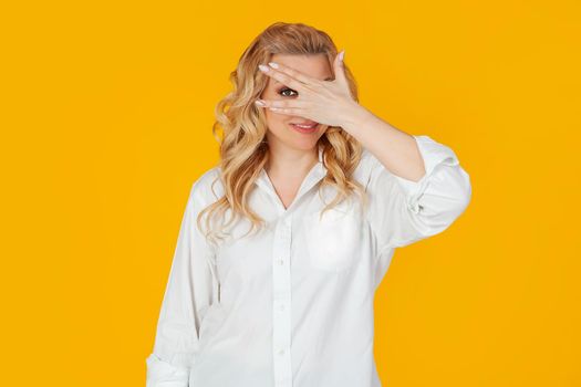Portrait of a middle-aged European blonde woman in a white shirt, closes her eyes with her hands and looks through her fingers, smiling. Standing on a yellow background. A look into the future