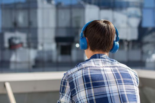 Back view of young man in checkered shirt with wireless headphones looking at cityscape. Guy listening to music on headphones in front of modern building