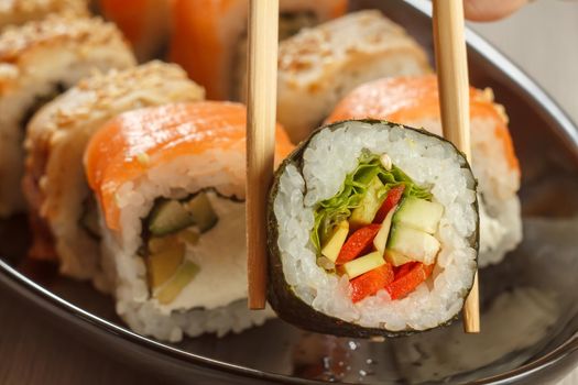 Two chopsticks holding Hosomaki with vegetables and different sushi rolls with seafood on ceramic plate on the background. Japanese cuisine. Shallow depth of field. Focus on roll with chopsticks