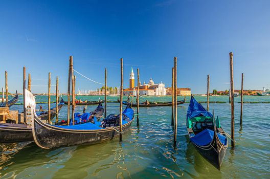 Gondolas moored docked on water in Venice. Gondoliers sailing San Marco basin waterway. San Giorgio Maggiore island with Campanile San Giorgio in Venetian Lagoon, blue clear sky, Veneto Region, Italy