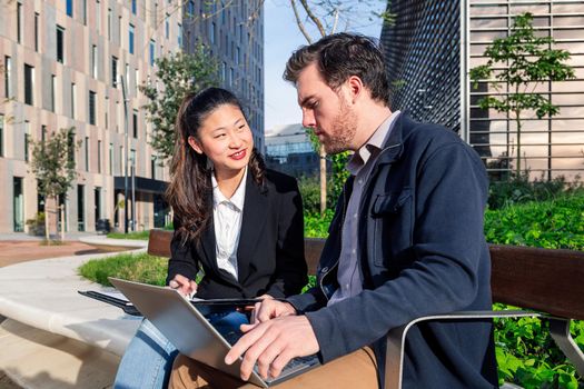 asian woman and caucasian man working together with laptop in a park next to the office, concept of coworkers and diversity at work