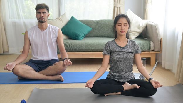 Young couple in sports clothes is meditating ,sitting in lotus position on mat while doing yoga at home. Sport at home, workout, fitness and exercises.