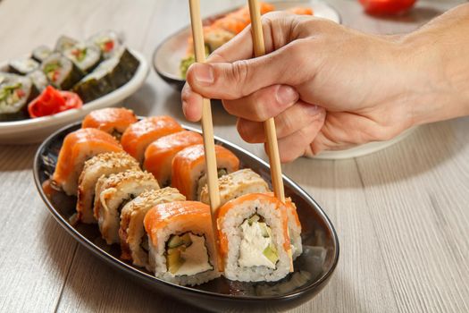 Male hand with two chopsticks holding Uramaki Philadelphia roll and different sushi rolls with seafood on ceramic plates on the background. Japanese cuisine. Focus on roll with chopsticks