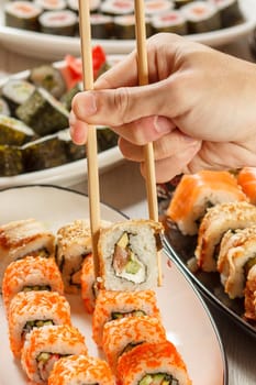 Male hand with two chopsticks holding Uramaki roll with Conger, California and different sushi rolls with seafood on ceramic plates on the background. Japanese cuisine. Focus on roll with chopsticks