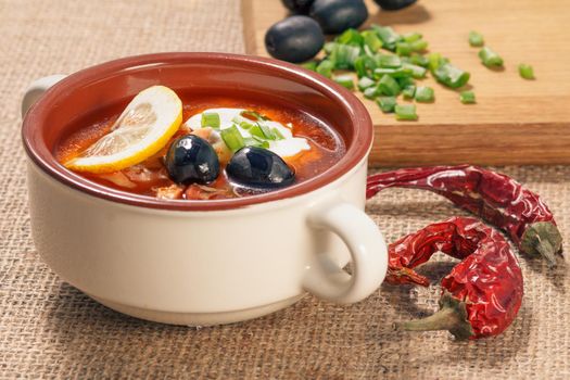 Soup saltwort with meat, smoked sausages, potatoes, tomatoes, marinated pickled cucumber, lemon, black olives and sour cream in ceramic soup bowl, dried red peppers, cutting board and ingredients on sackcloth