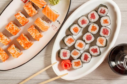 Different sushi rolls with rice, vegetable, seafood on ceramic plates and glass bottle with soy sauce on wooden desk. Top view