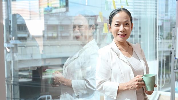 Confident middle aged business leader standing near large window at office with city buildings in background.