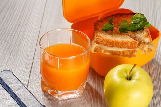 Yellow sandwich box with toasted slices of bread, cheese and green parsley, green apple, glass of orange juice and notebook on wooden desk. School breakfast.