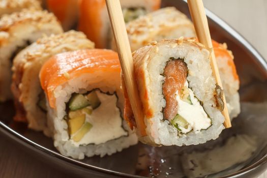 Two chopsticks holding Uramaki with Conger and different sushi rolls with seafood on ceramic plate on the background. Japanese cuisine. Shallow depth of field. Focus on roll with chopsticks