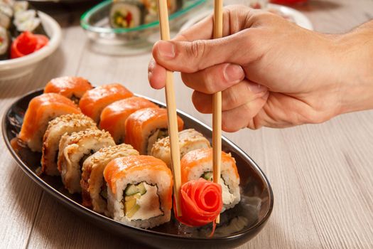 Male hand with two chopsticks holding pickled ginger in the form of red rose and different sushi rolls with rice, vegetable, seafood on ceramic plates on the background