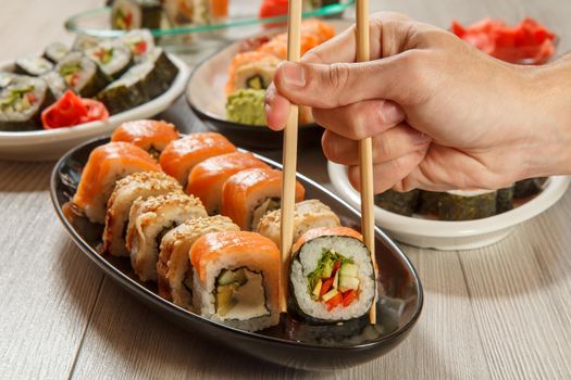 Male hand with two chopsticks holding Hosomaki roll with vegetables and different sushi rolls with seafood on ceramic plates on the background. Japanese cuisine. Focus on roll with chopsticks