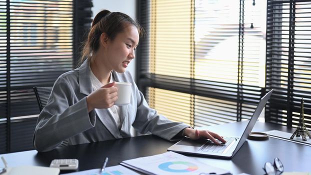 Professional chartered accountant drinking coffee and working with laptop computer at office desk.
