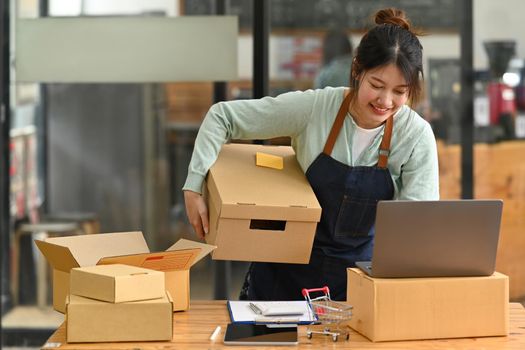 Startup small business owner checking or accepting orders on her laptop and preparing packages for delivery. Online selling, E-commerce.