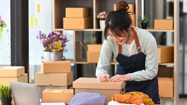 Happy young woman online seller writing address on cardboard box while preparing boxes of product for delivery. Online selling, E-commerce, Online Shopping.