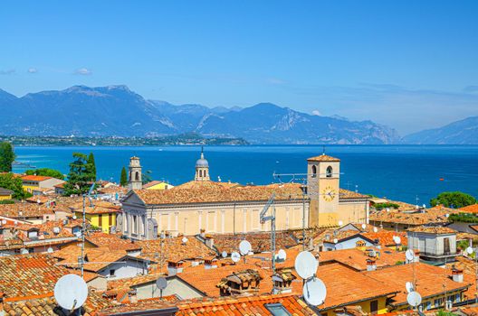 Aerial view of Desenzano del Garda town with bell tower of Duomo di Santa Maria Maddalena Cathedral church, red tiled roof buildings, Garda Lake water surface, mountain range, Lombardy, Northern Italy