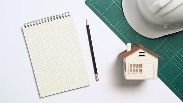 Above view blank notebook, house model and safety helmet on white table.