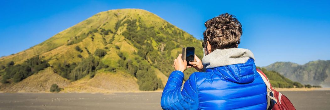 BANNER, LONG FORMAT Young man tourist takes a photo of Batok volcano on his cellphone. He is in the Bromo Tengger Semeru National Park on the Java Island, Indonesia. He enjoys magnificent view on the Bromo or Gunung Bromo on Indonesian, Semeru and other volcanoes located inside of the Sea of Sand within the Tengger Caldera. One of the most famous volcanic objects in the world. Travel to Indonesia concept.