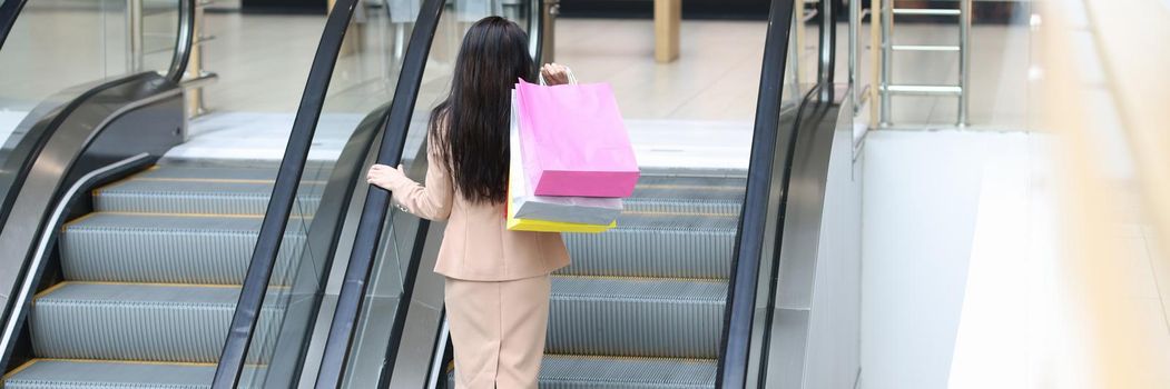 Beautiful woman with packages climbs the escalator of the mall, view from the back. Shopping pleasure, search for discounted items