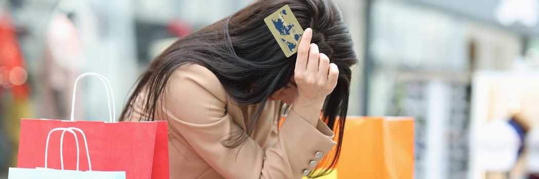 A young woman sits in a shopping center with a credit card and a phone in her hands. Checking the balance after buying