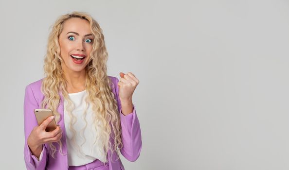 Happy triumphant woman holds a smartphone, fist and an enthusiastic smile, receives great fantastic news using a text message on her mobile phone. Wins the online lottery, receives the first prize.