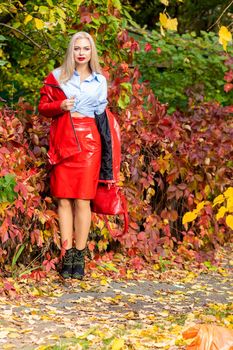 A fashionable woman wears a trendy white and blue shirt and red skirt and leather jacket on a city street. Stylish young blonde lady with bright makeup.