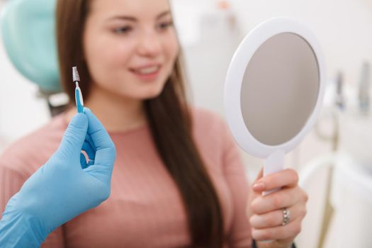 Unrecognizable dentist holding interdental toothbrush, female patient checking her teeth in the mirror on background