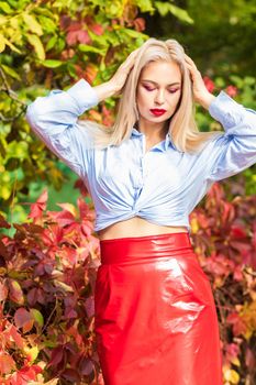 A fashionable woman wears a trendy white and blue shirt and red skirt and leather jacket on a city street. Stylish young blonde lady with bright makeup.