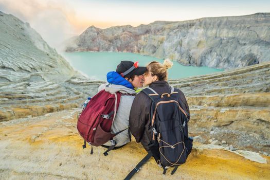 Young tourist man and woman sit at the edge of the crater of the Ijen volcano or Kawah Ijen on the Indonesian language. Famous volcano containing the biggest in the world acid lake and sulfur mining spot at the place where volcanic gasses come from the volcano. They enjoy the magnificent view on the crater.