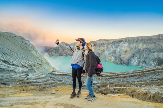 Young tourist man and woman stand at the edge of the crater of the Ijen volcano or Kawah Ijen on the Indonesian language. Famous volcano containing the biggest in the world acid lake and sulfur mining spot at the place where volcanic gasses come from the volcano. They wear respirators to protect from dangerous volcanic gasses.