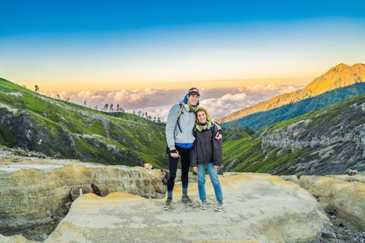 Young tourist man and woman stand at the edge of the crater of the Ijen volcano or Kawah Ijen on the Indonesian language. Famous volcano containing the biggest in the world acid lake and sulfur mining spot at the place where volcanic gasses come from the volcano. They wear respirators to protect from dangerous volcanic gasses.