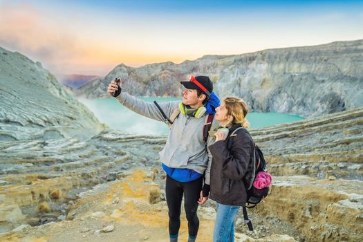 Young tourist man and woman stand at the edge of the crater of the Ijen volcano or Kawah Ijen on the Indonesian language. Famous volcano containing the biggest in the world acid lake and sulfur mining spot at the place where volcanic gasses come from the volcano. They wear respirators to protect from dangerous volcanic gasses.