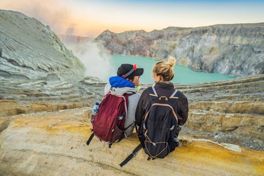 Young tourist man and woman sit at the edge of the crater of the Ijen volcano or Kawah Ijen on the Indonesian language. Famous volcano containing the biggest in the world acid lake and sulfur mining spot at the place where volcanic gasses come from the volcano. They enjoy the magnificent view on the crater.