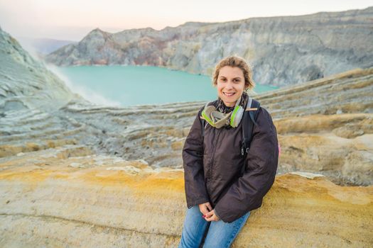 Young woman tourist sitting at the edge of the crater of the Ijen volcano or Kawah Ijen on the Indonesian language. Famous volcano containing the biggest in the world acid lake and sulfur mining spot at the place where volcanic gasses come from the volcano. She wears a respirator to protect from dangerous volcanic gasses.