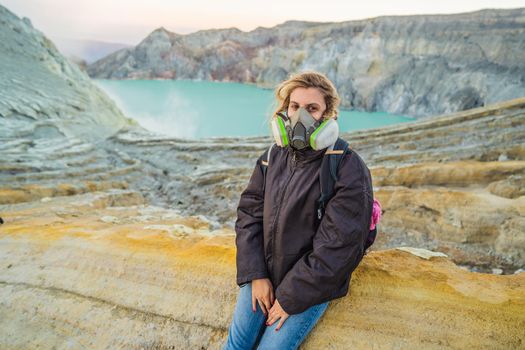 Young woman tourist sitting at the edge of the crater of the Ijen volcano or Kawah Ijen on the Indonesian language. Famous volcano containing the biggest in the world acid lake and sulfur mining spot at the place where volcanic gasses come from the volcano. She wears a respirator to protect from dangerous volcanic gasses.