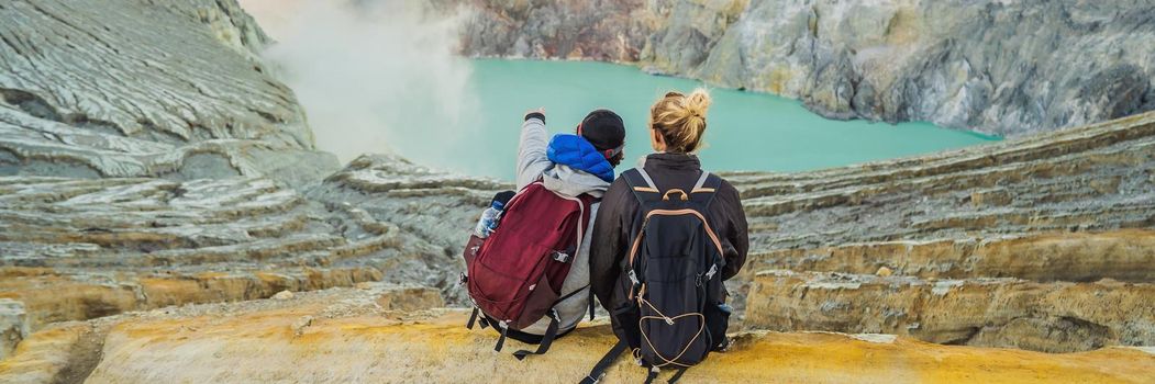 BANNER, LONG FORMAT Young tourist man and woman sit at the edge of the crater of the Ijen volcano or Kawah Ijen on the Indonesian language. Famous volcano containing the biggest in the world acid lake and sulfur mining spot at the place where volcanic gasses come from the volcano. They enjoy the magnificent view on the crater.