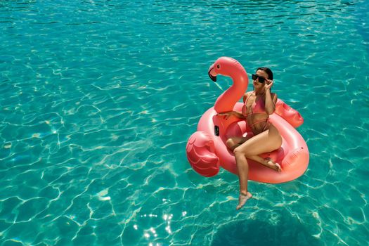 Above view of brunette girl swimming in pool, sitting on pink flamingo. Slim young female in swimsuit and sunglasses relaxing, sunbathing, enjoying. Concept of summertime.