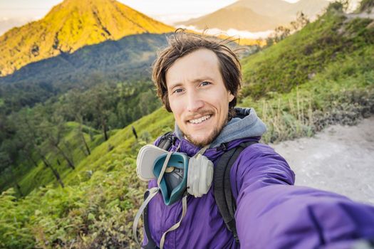Young man tourist makes a selfie with the magnificent views on green mountains from a mountain road trecking to the Ijen volcano or Kawah Ijen on the Indonesian language. Famous volcano containing the biggest in the world acid lake and sulfur mining spot at the place where volcanic gasses come from the volcano. Tourists hike this road to meet the sunrise at the volcano.