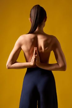 Back view of flexible girl with bare back standing backwards with prayer hands indoors. Brunette with pony tail stretching, meditating, relaxing. Isolated onn yellow studio background.
