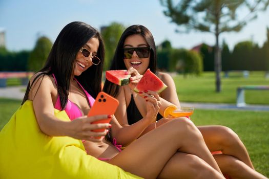 Front view of two pretty brunette women sitting on armchair bags, taking selfie. Seductive young women relaxing, smiling, talking, eating watermelon. Concept of summertime.